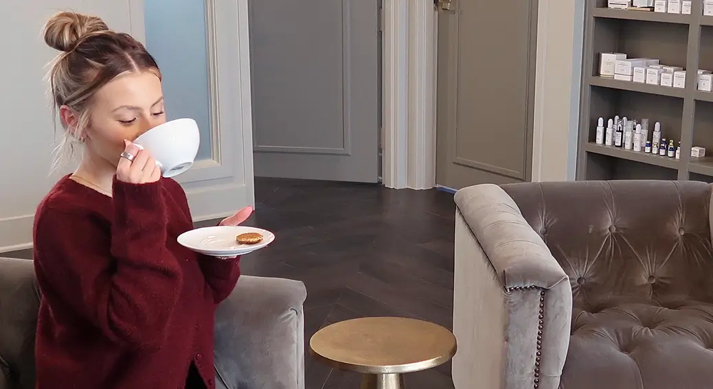 Woman in a burgundy sweater sitting on a gray chair drinking from a white cup and holding a plate with a cookie.