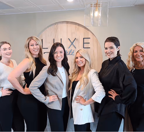 Six women standing indoors in front of a round wooden wall sign that reads 'LUXE beauté', all posing with hands on hips and smiling.