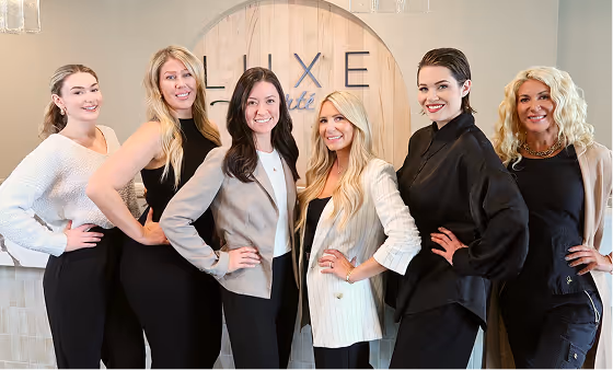 Six professional women standing side by side in a modern office with a wooden Luxe sign in the background.