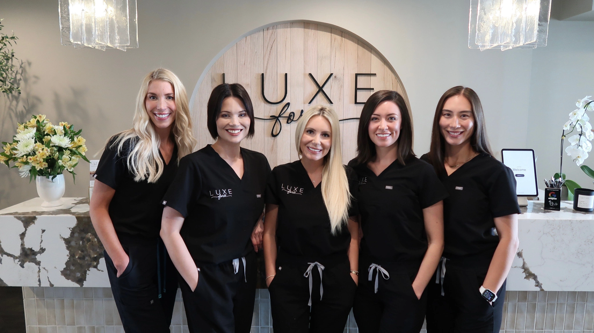 Six professional women standing side by side in a modern office with a wooden Luxe sign in the background.