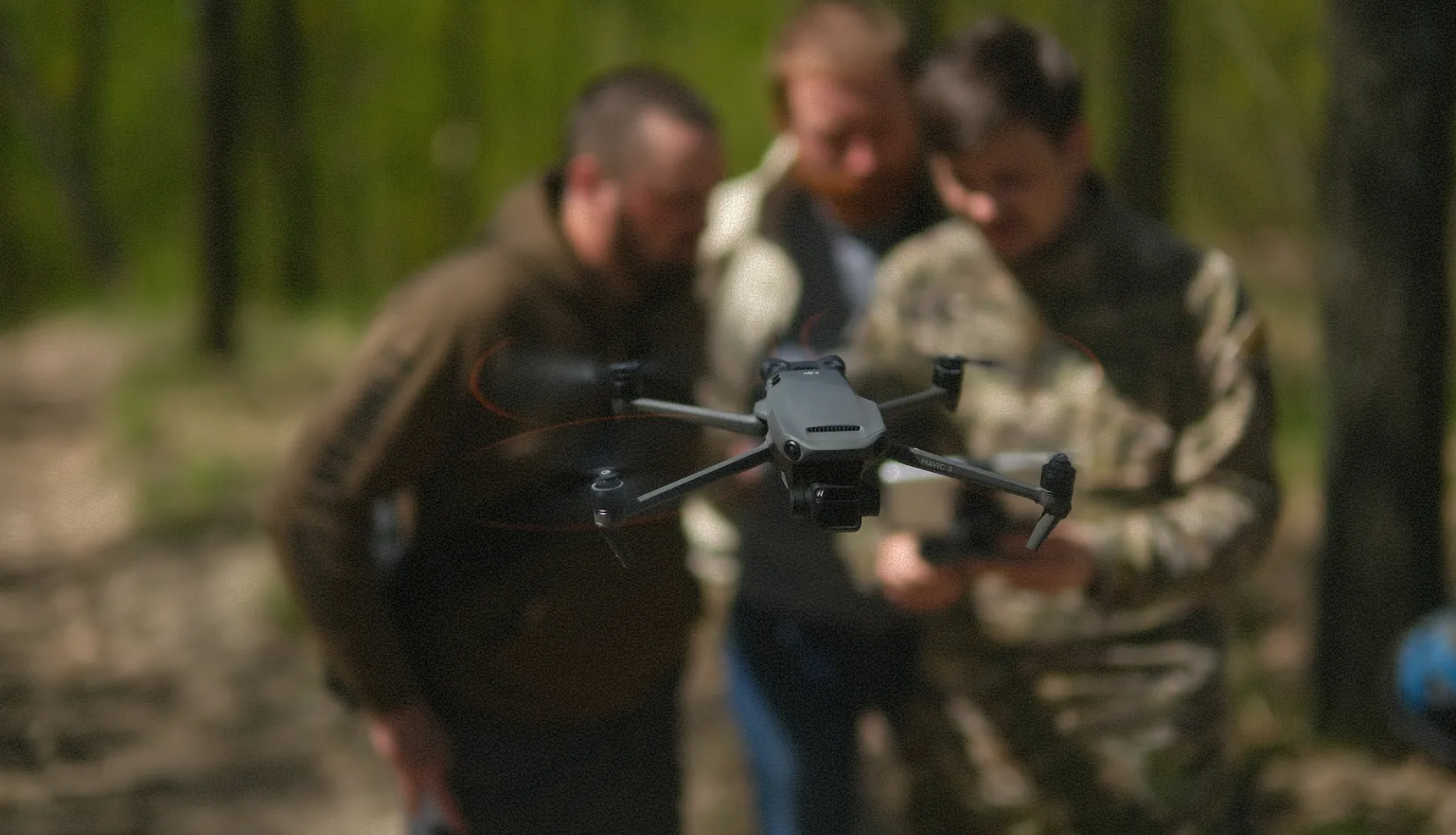 Dron latający na pierwszym planie, a za nim trzech mężczyzn stojących na zewnątrz w lesie i skupionych na kontrolerze drona.