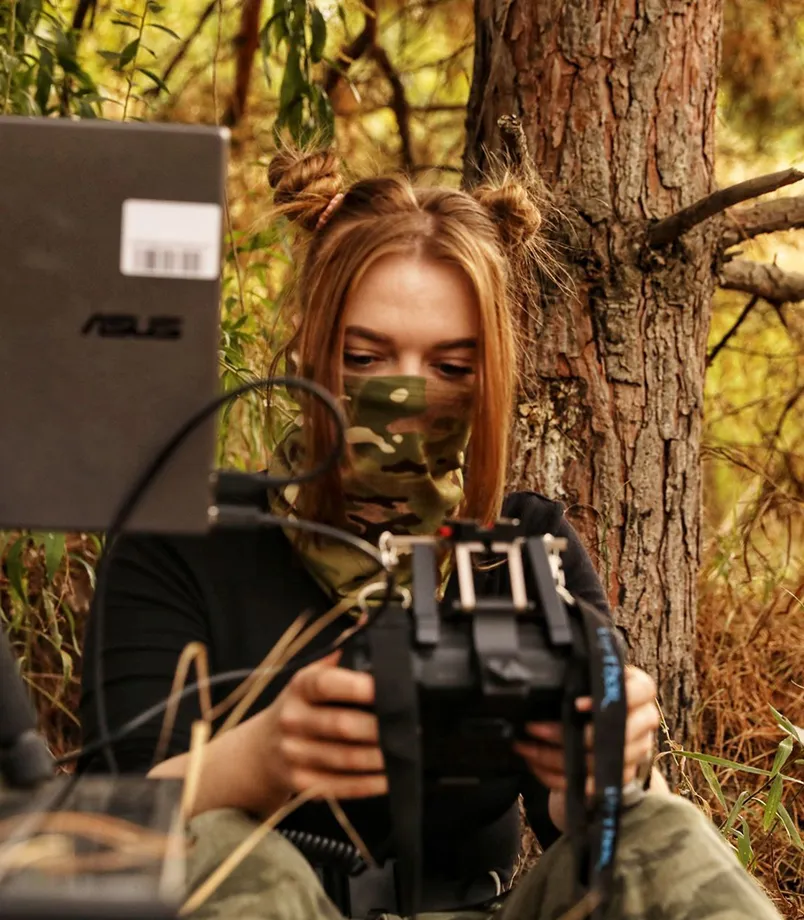 Młoda kobieta w moro szaliku na twarzy obsługująca urządzenie elektroniczne na tle drzewa w lesie.