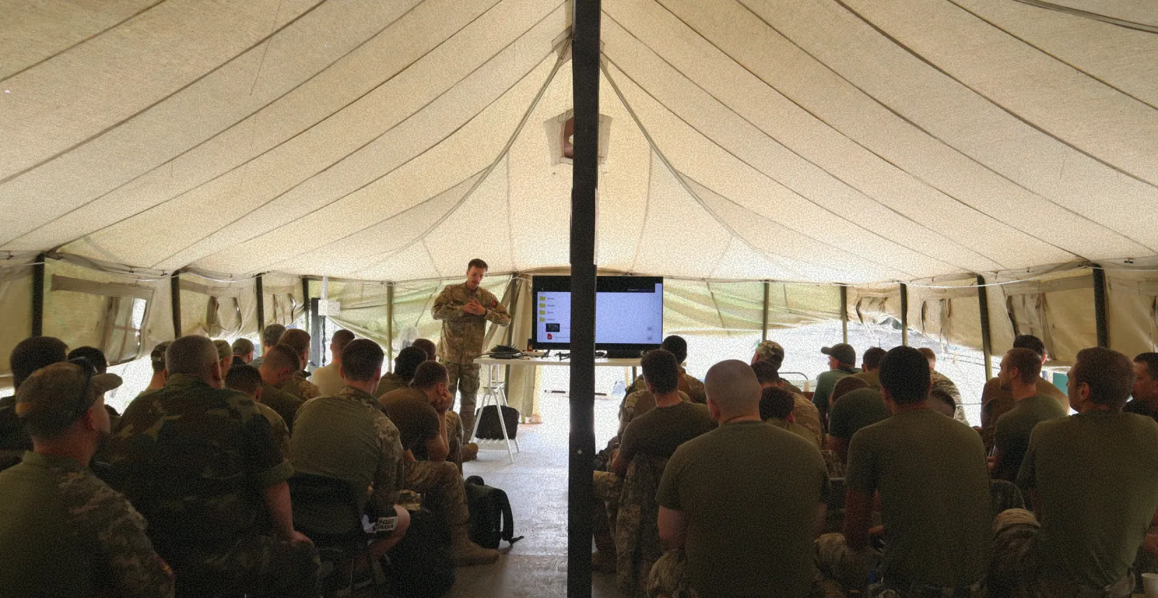Żołnierz prowadzi prezentację na telewizorze wewnątrz dużego namiotu przed grupą żołnierzy siedzących na krzesłach.