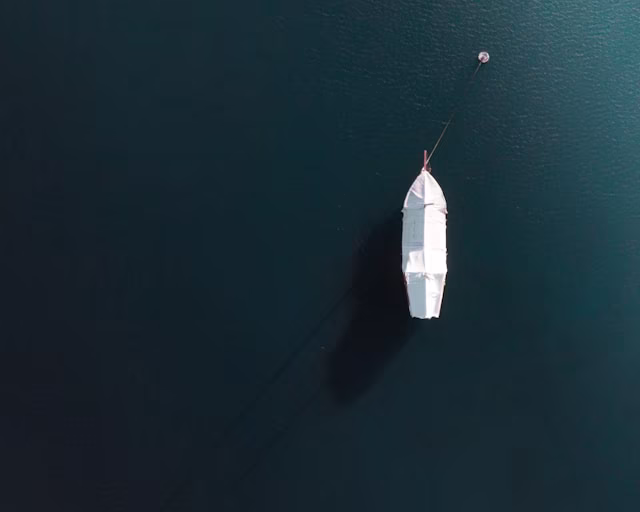 Top-down view of a small white boat anchored in calm dark water casting a shadow.