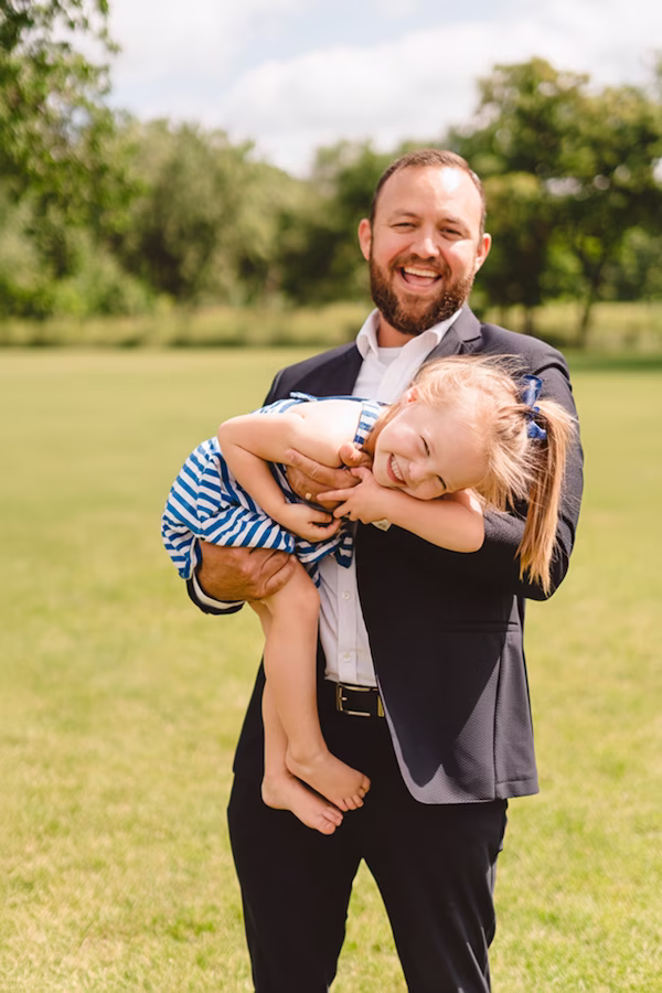 Robert Lopez, CFP® smiling in suit holding happy young girl in blue striped dress in a green outdoor setting.