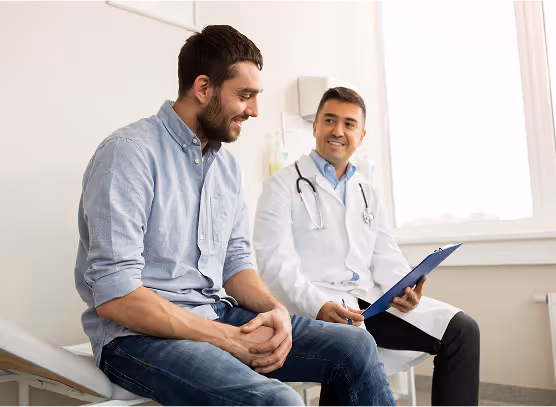 Male doctor in white coat consulting a smiling male patient sitting on an examination table in a bright clinic room.
