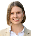 Smiling woman with shoulder-length brown hair wearing a beige blazer and white collared shirt against a white background.