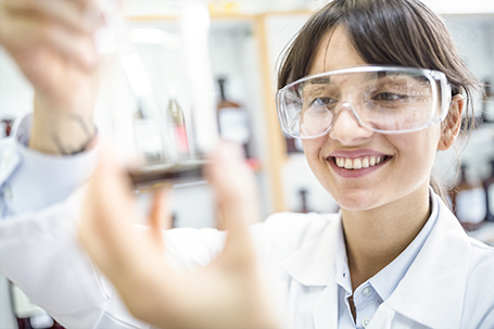 Smiling female scientist wearing safety goggles and a lab coat examining a glass beaker in a laboratory.