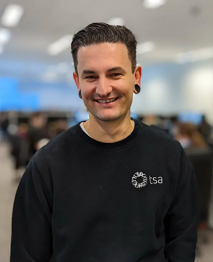 Smiling man with short dark hair and large black ear gauges wearing a black TSA sweatshirt in an office setting.