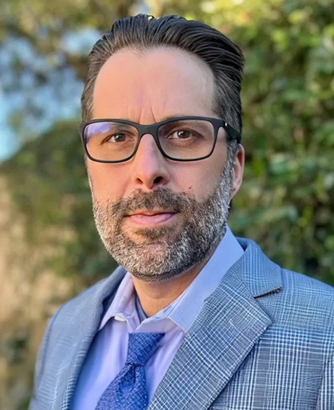 Man with dark hair, beard, and glasses wearing a light blue shirt, blue patterned tie, and gray plaid blazer outdoors with green foliage background.