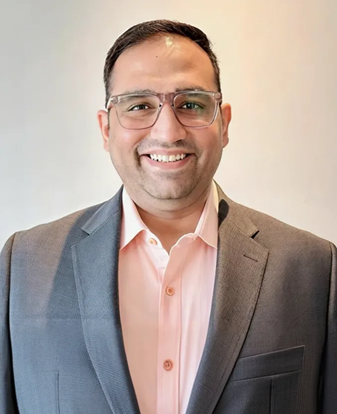 Smiling man wearing glasses, a light pink shirt, and a gray blazer against a plain light background.