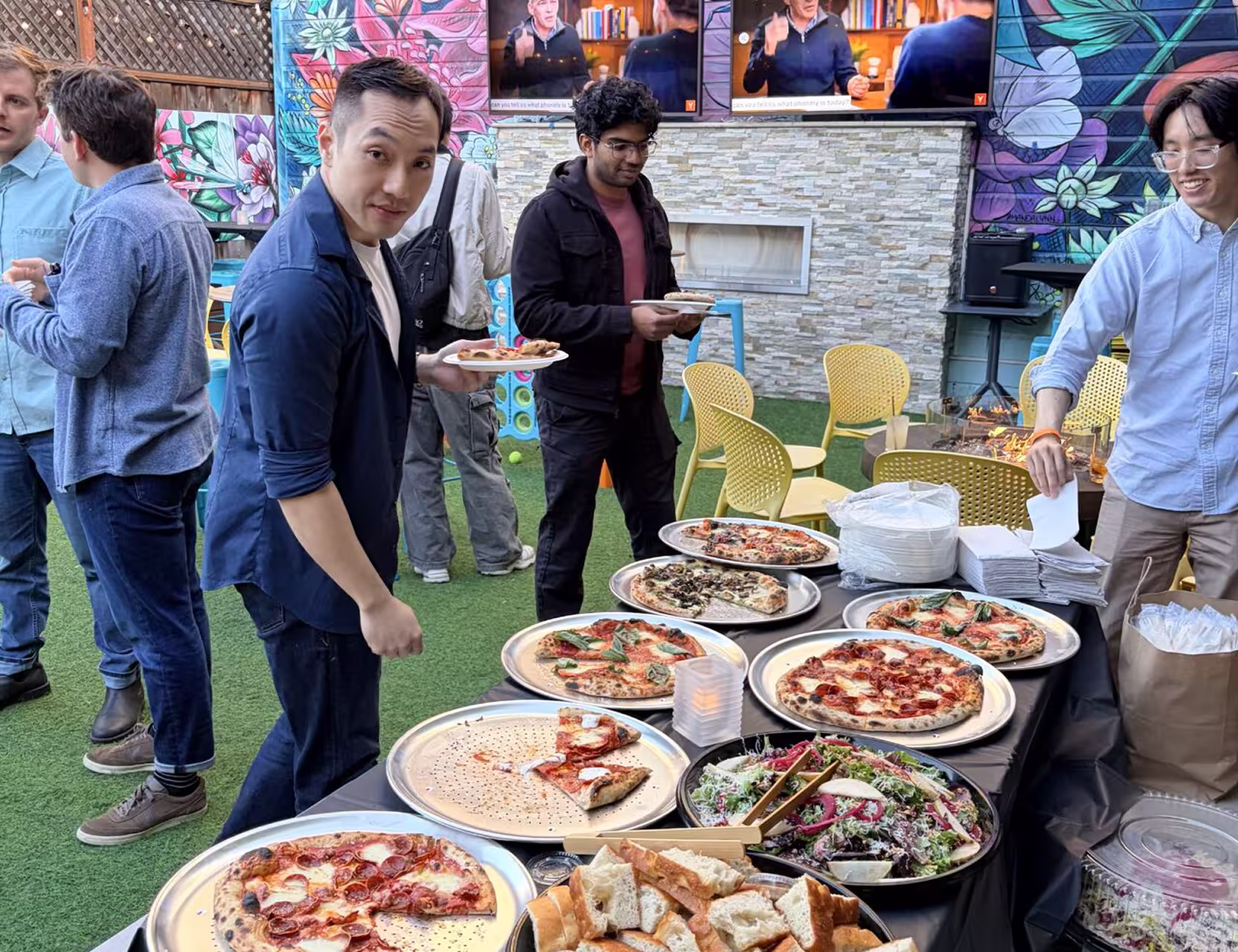 People serving themselves pizza and salad at an outdoor party with colorful mural background.