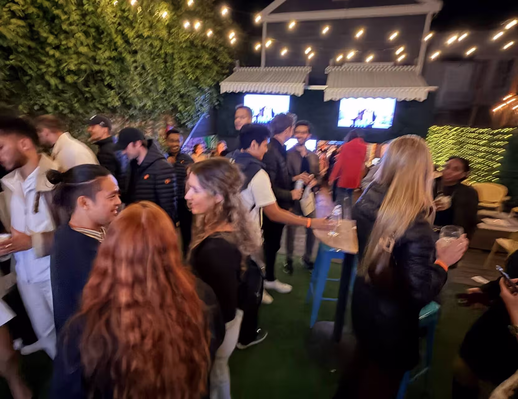 Group of people socializing under string lights at an outdoor gathering with screens in the background.