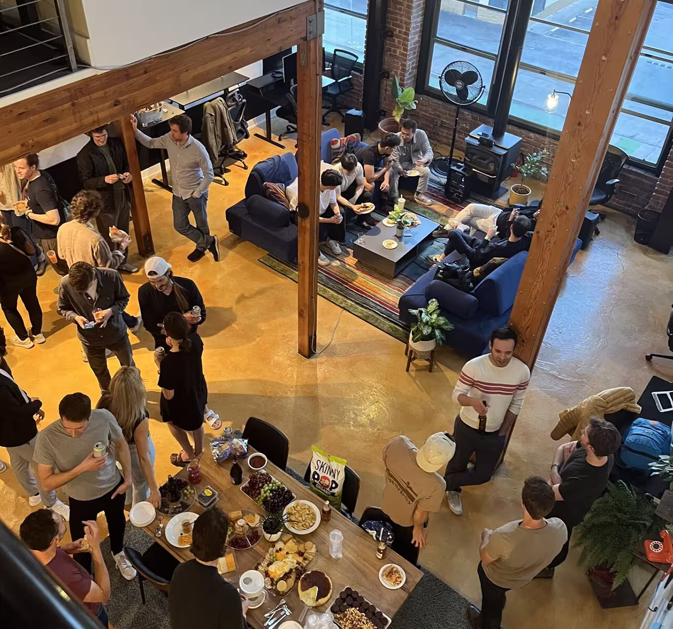 Group of people socializing and eating snacks in a spacious, modern loft with wooden beams.