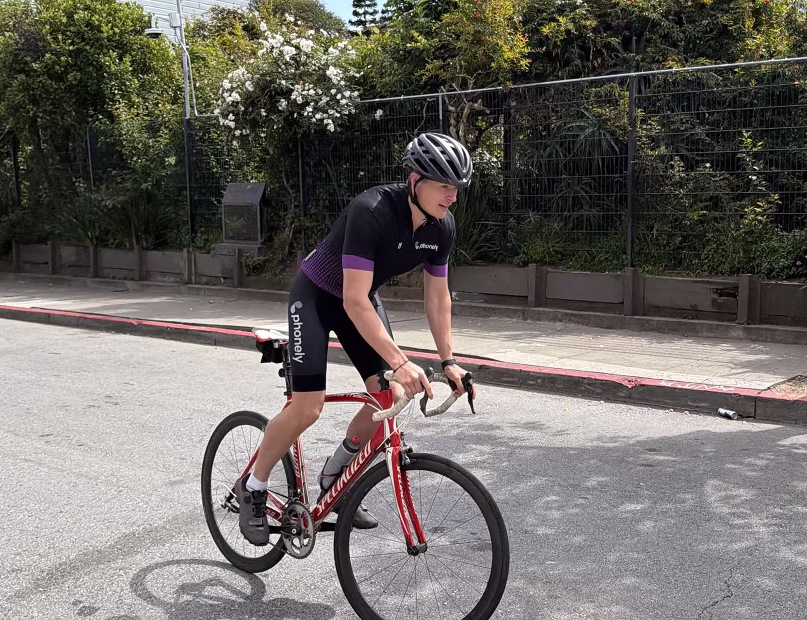 Cyclist wearing black helmet and Phonely outfit riding a red Specialized road bike on a street.