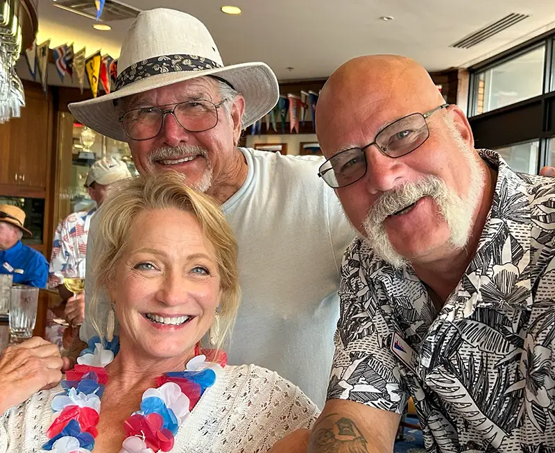 Three smiling adults posing closely together indoors, with one woman wearing a red, white, and blue lei and two men wearing glasses.