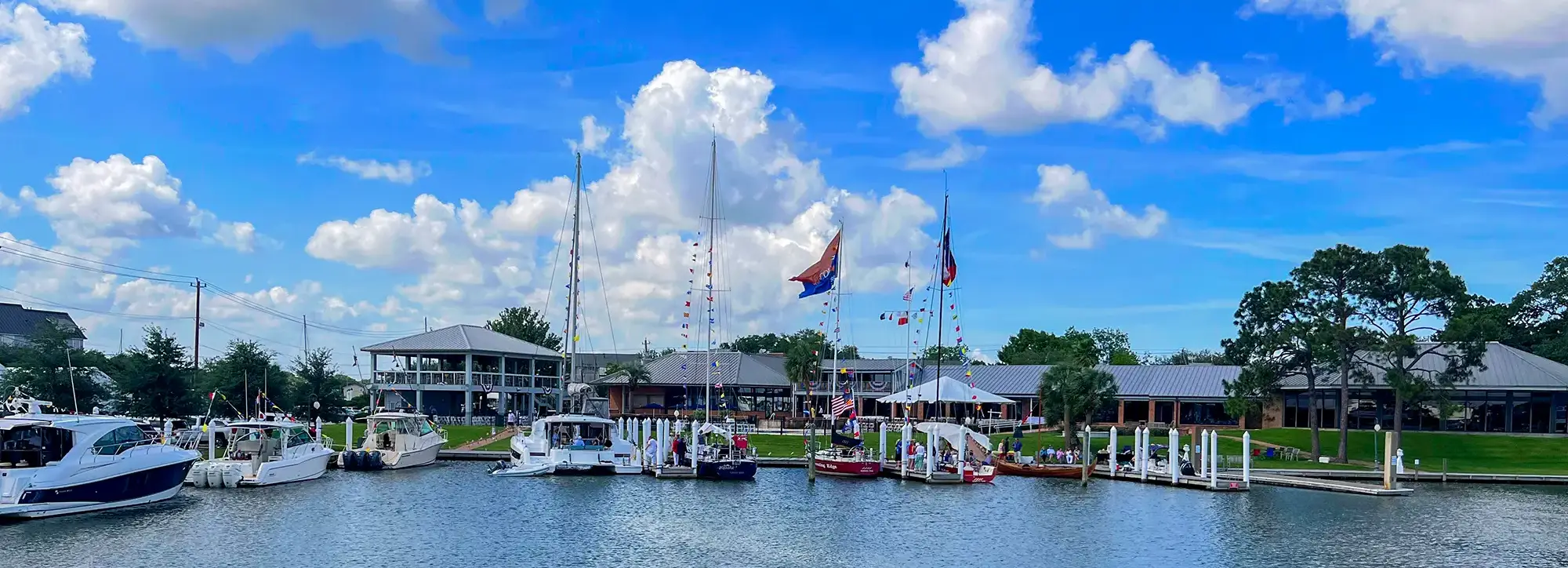 Boats docked at a marina with a building, green lawn, and blue sky with scattered clouds in the background.