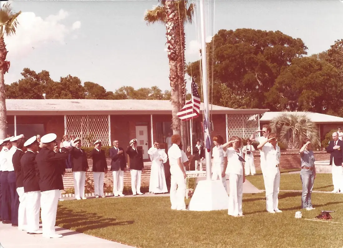 Group of people, including uniformed personnel and civilians, saluting in front of two raised flags on a sunny day outside a building.