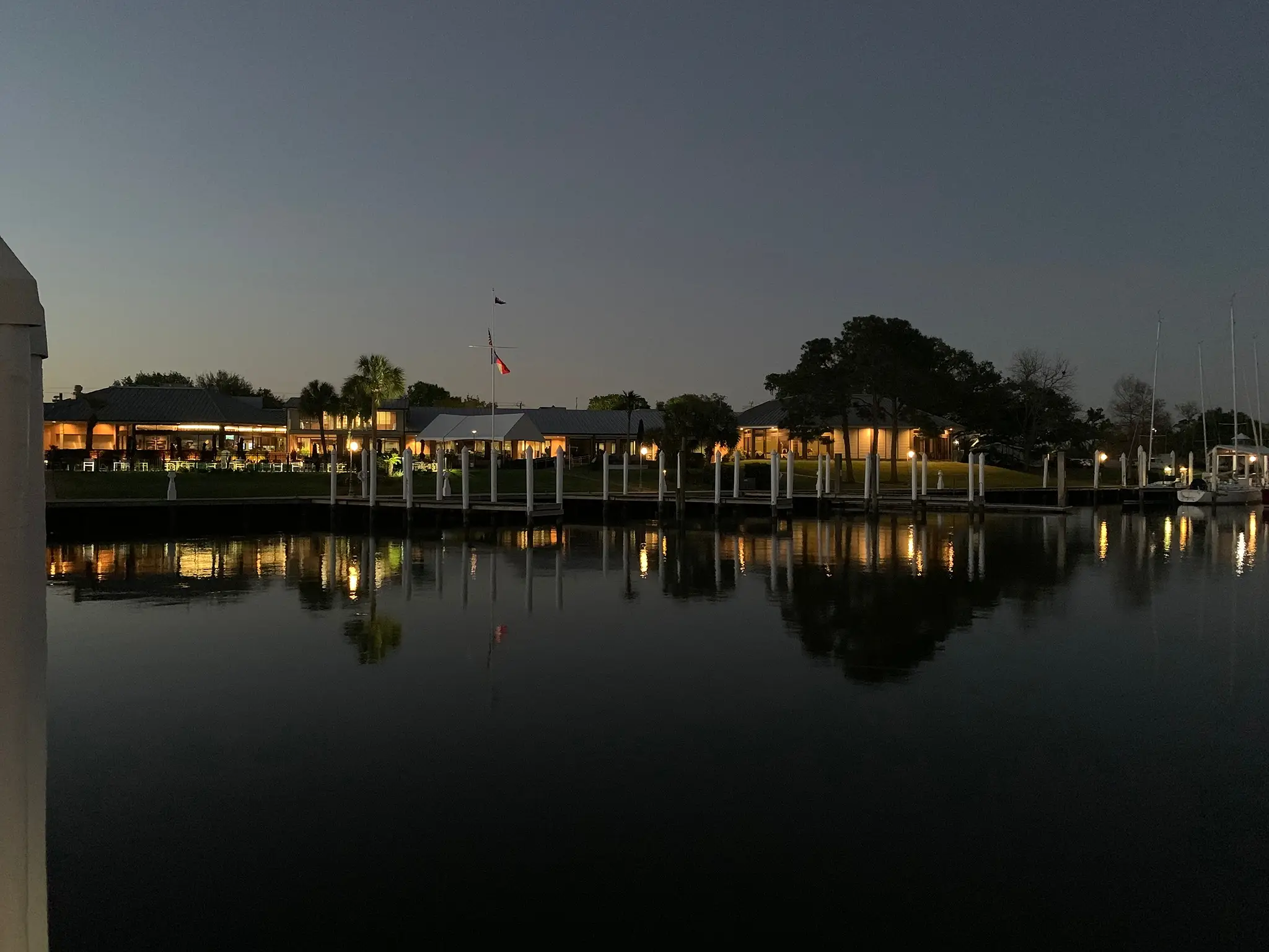 Evening view of a marina with dock posts, sailboats, and illuminated buildings reflecting on calm water.