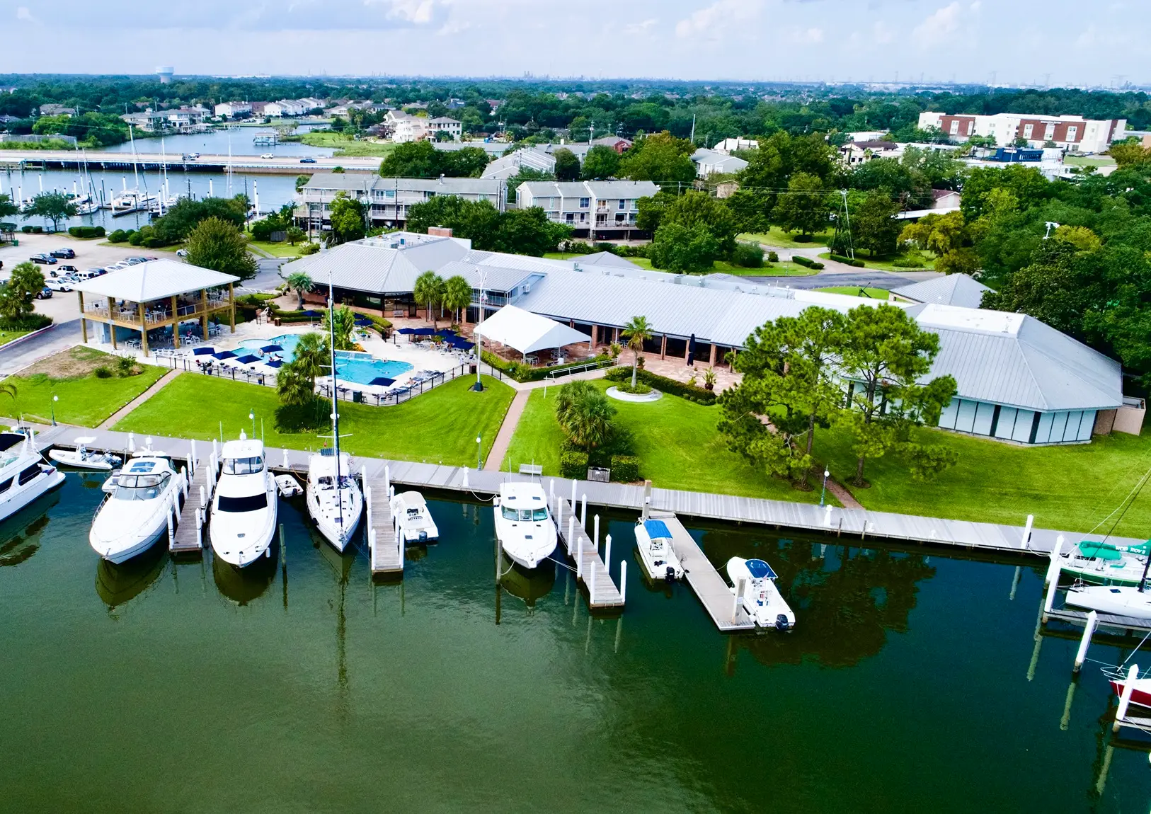Aerial view of a marina with several boats docked, a pool area, and a large building surrounded by green lawn and trees.