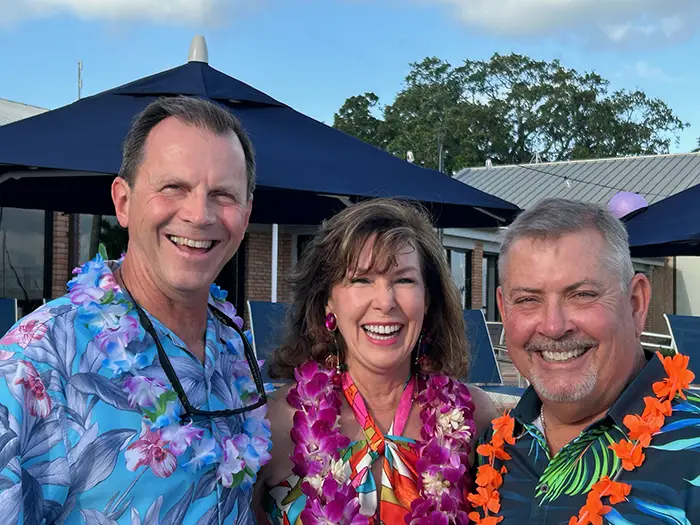 Three smiling adults wearing floral leis and colorful clothing at an outdoor gathering with umbrellas and buildings in the background.