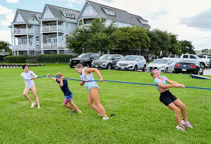 Four children playing tug-of-war on a grassy field with cars and houses in the background.