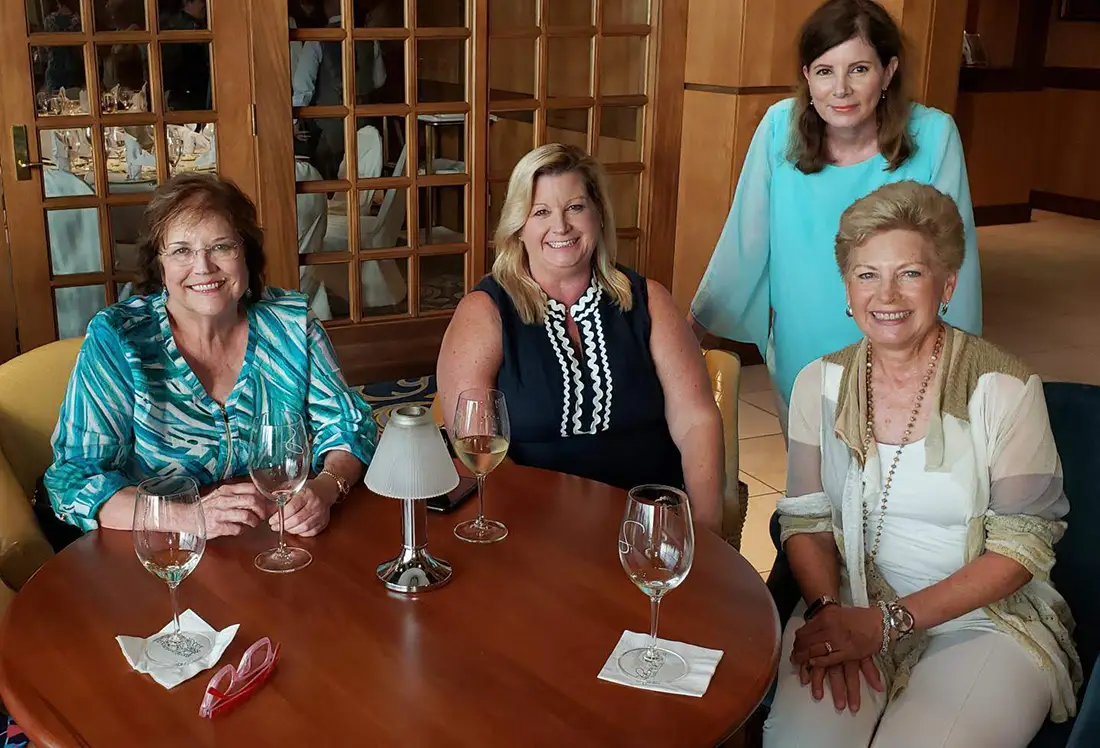 Four women smiling at a round wooden table with wine glasses and a small lamp in a warmly lit room.