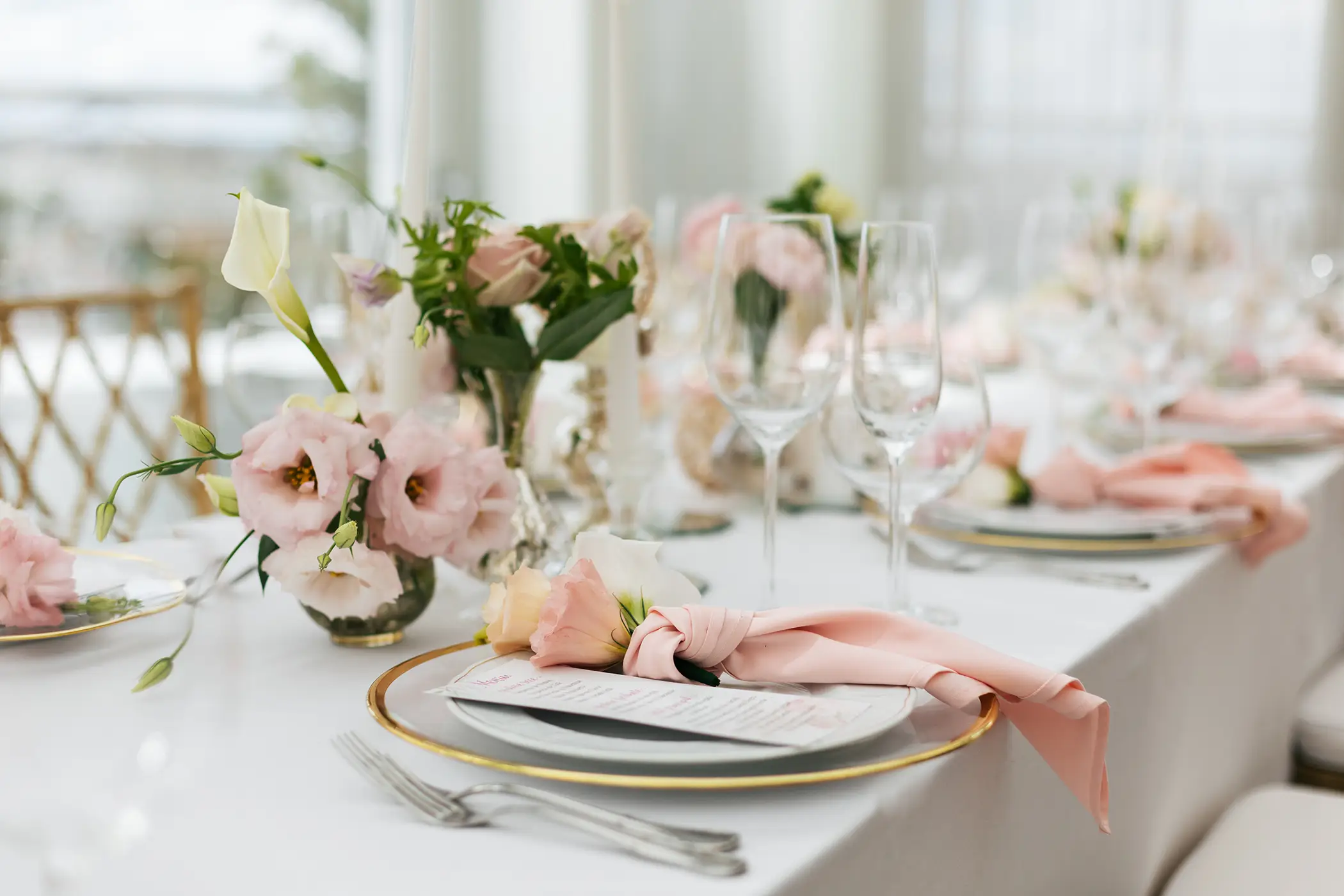 Elegant table setting with gold-rimmed plates, pink cloth napkins tied with flowers, and floral centerpieces on a white tablecloth.