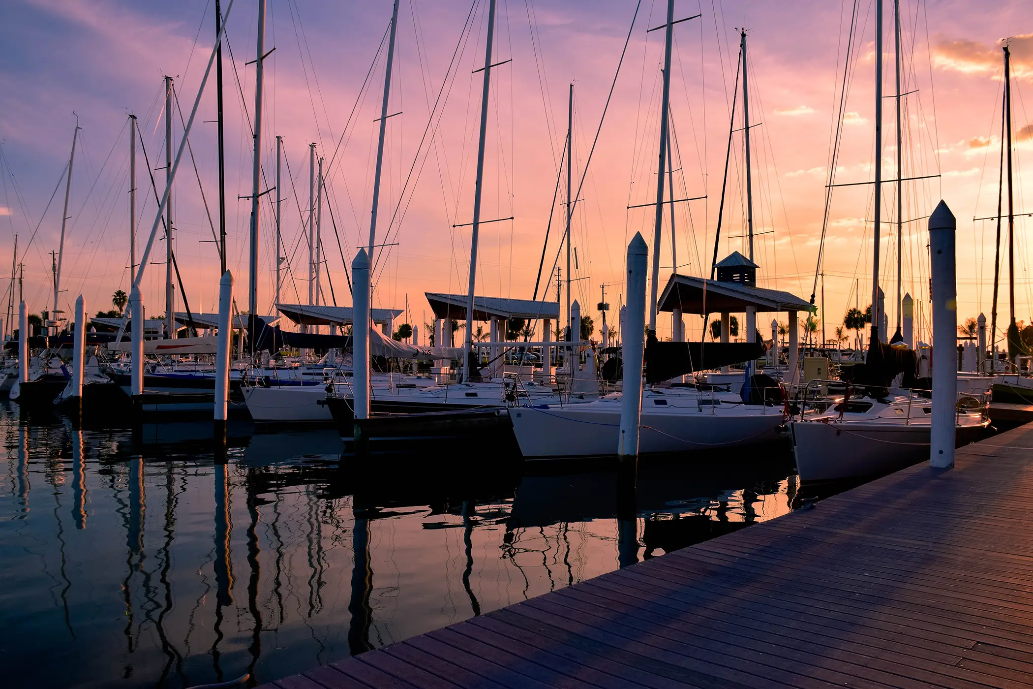 Sailboats docked at a marina during a colorful sunset with reflections on calm water.