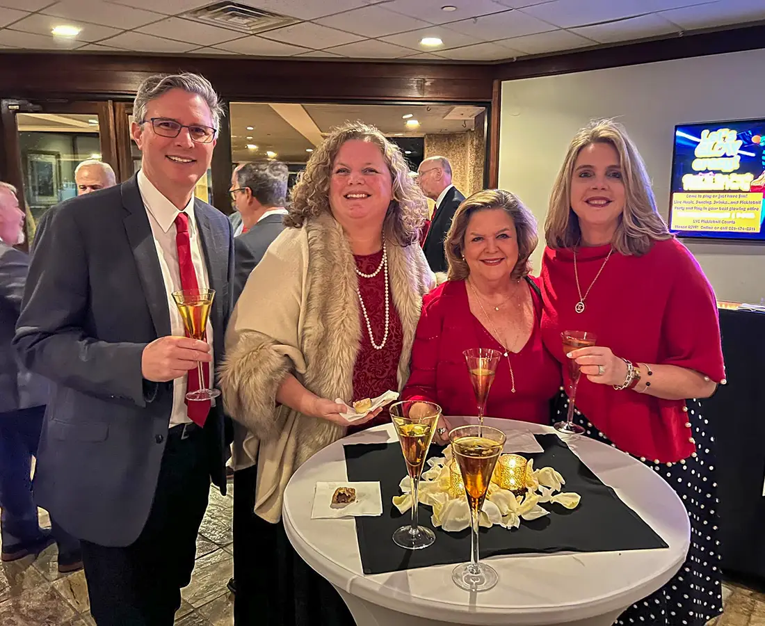 Four adults smiling and holding drinks around a table decorated with rose petals and candles at an indoor event.