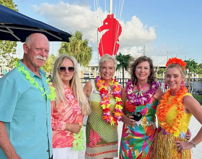 Five adults wearing colorful tropical clothing and flower leis posing outdoors near water and seahorse sculpture.