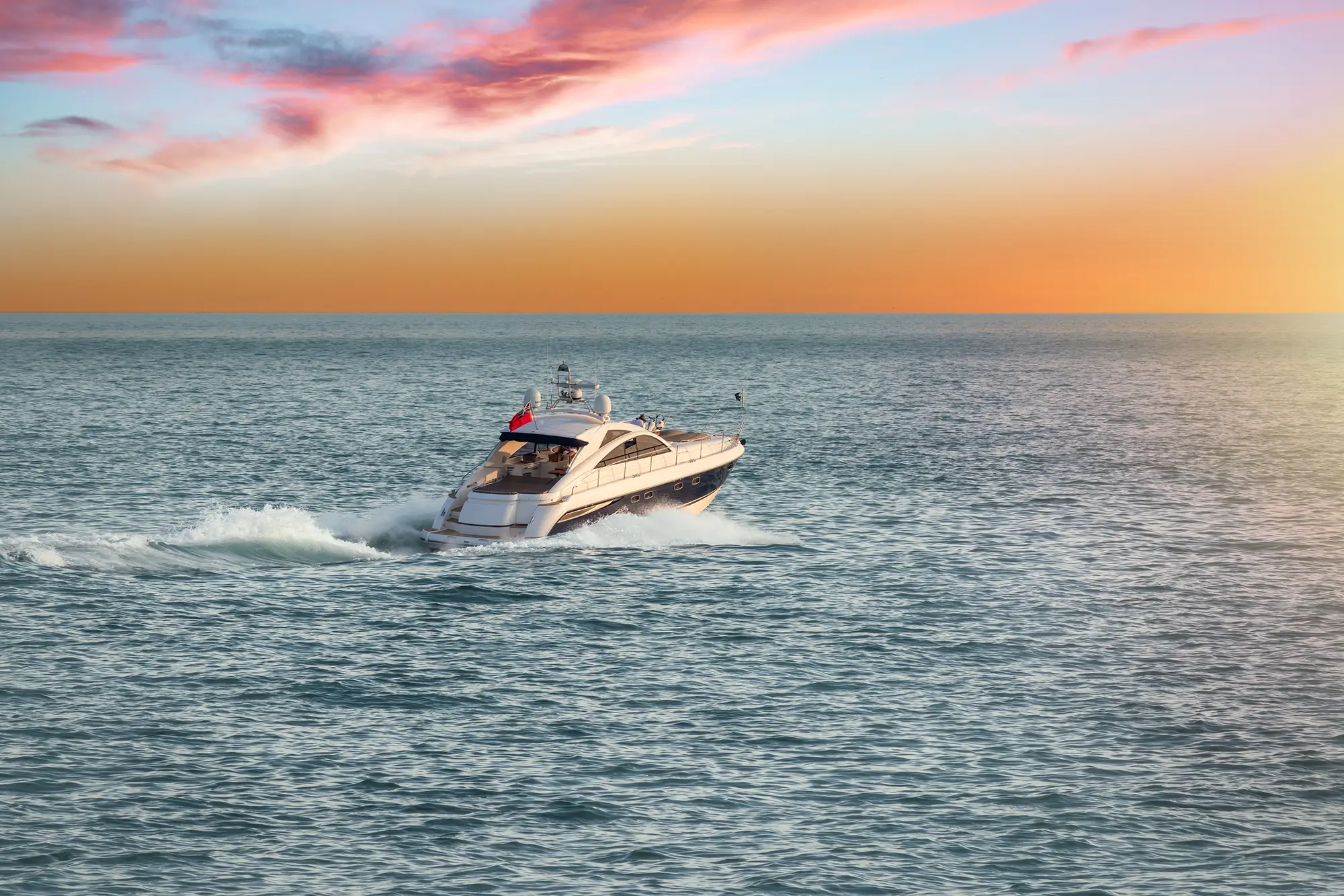White and navy yacht cruising on calm sea at sunset with pink and orange sky.