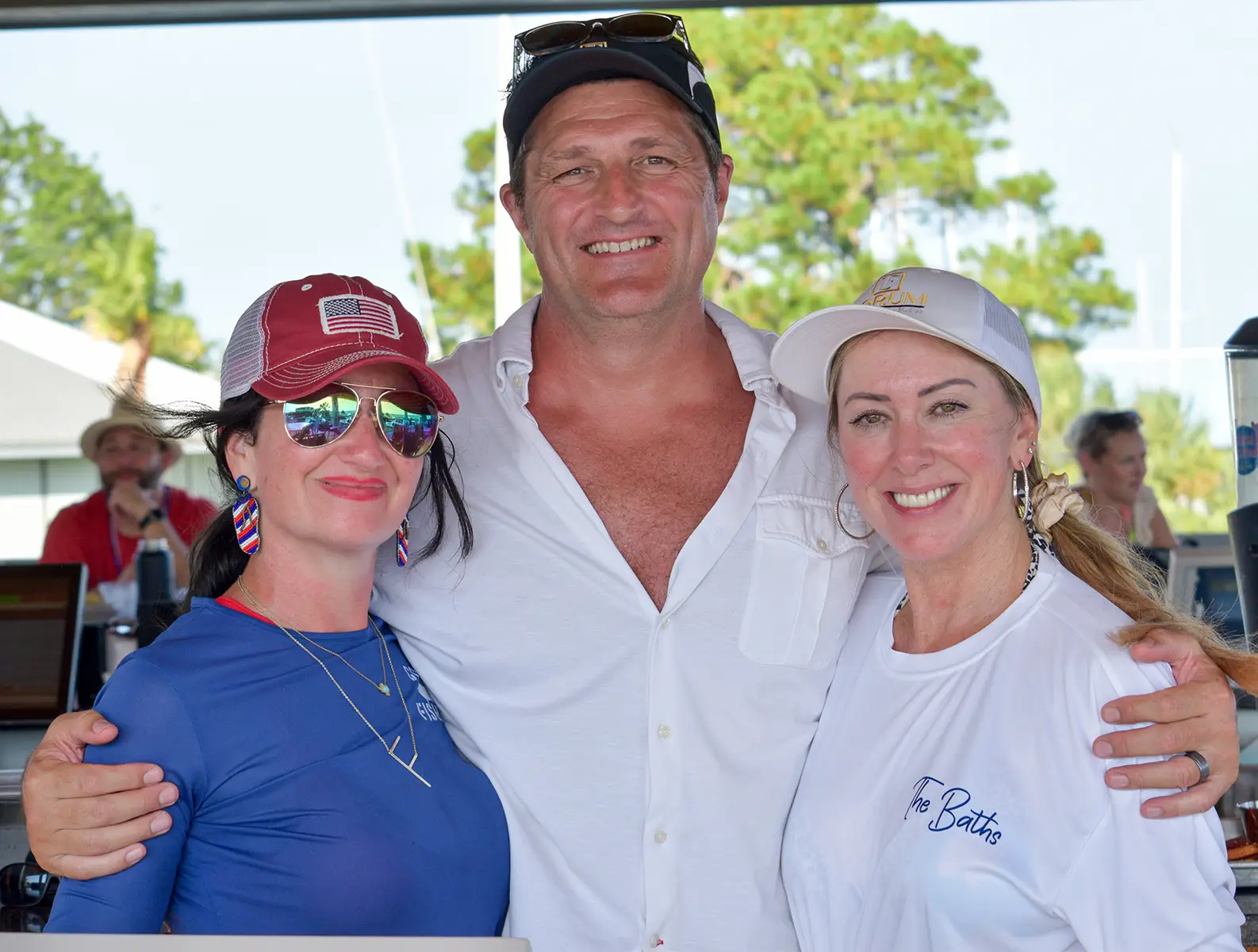 Three people smiling with arms around each other; woman in blue with American flag hat and sunglasses, man in white shirt and cap, woman in white shirt and cap.