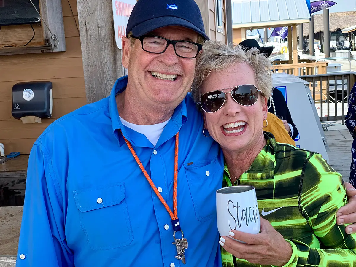 Smiling man in a blue shirt and cap embraces smiling woman in green patterned jacket holding a cup labeled 'Stacie'.