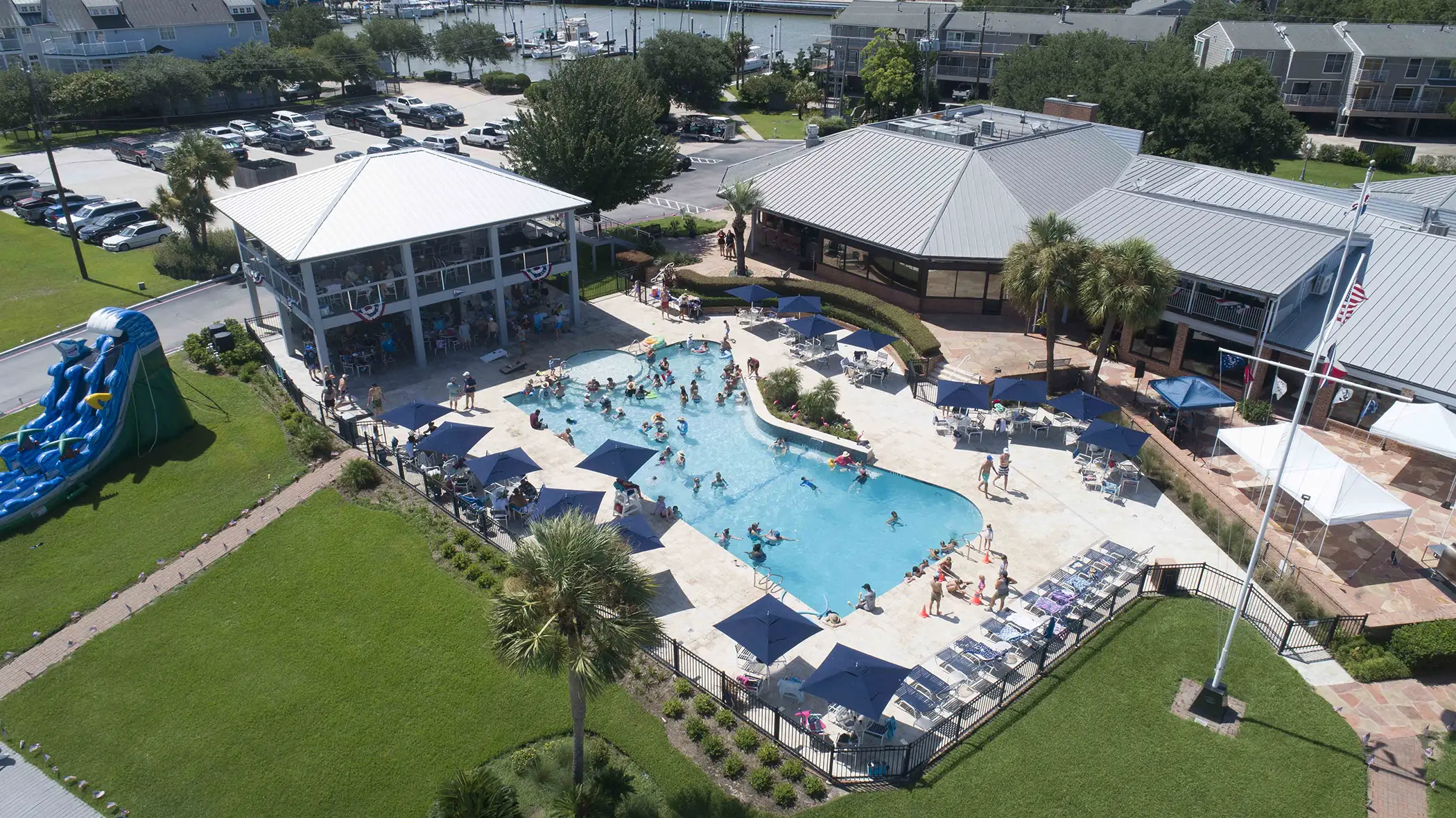 Aerial view of a busy outdoor swimming pool with people swimming and lounging under umbrellas, adjacent to buildings and a large grassy area with an inflatable water slide.