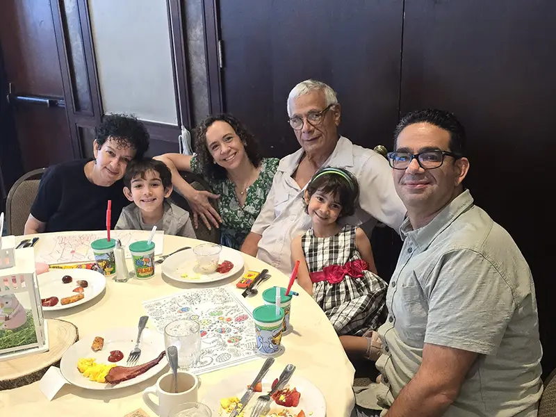 Six family members sitting closely around a dining table with plates of food and children's cups, smiling at the camera.