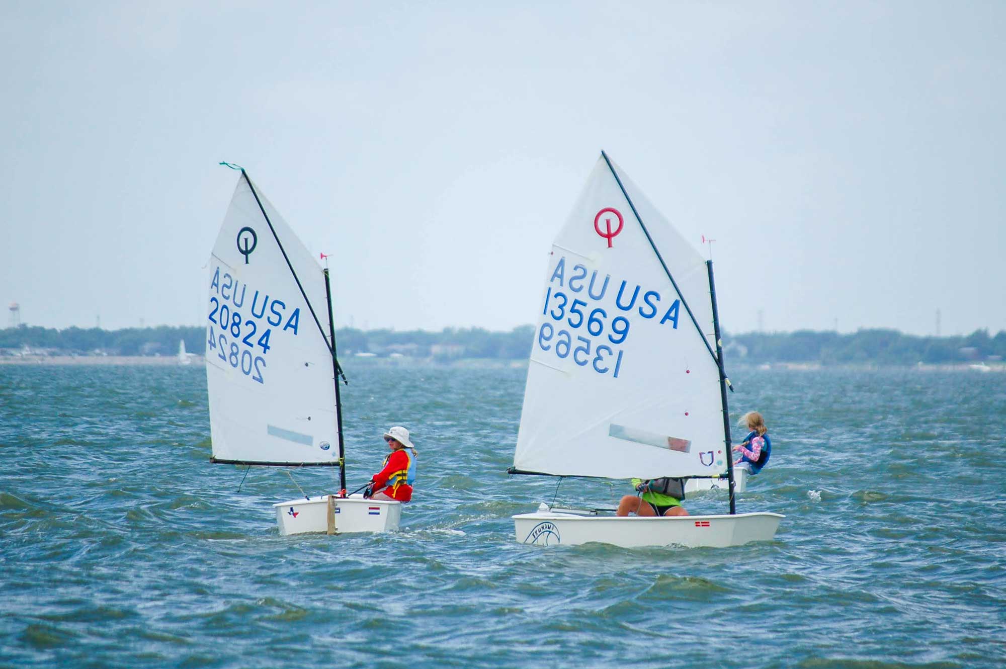Evening view of a marina with dock posts, sailboats, and illuminated buildings reflecting on calm water.