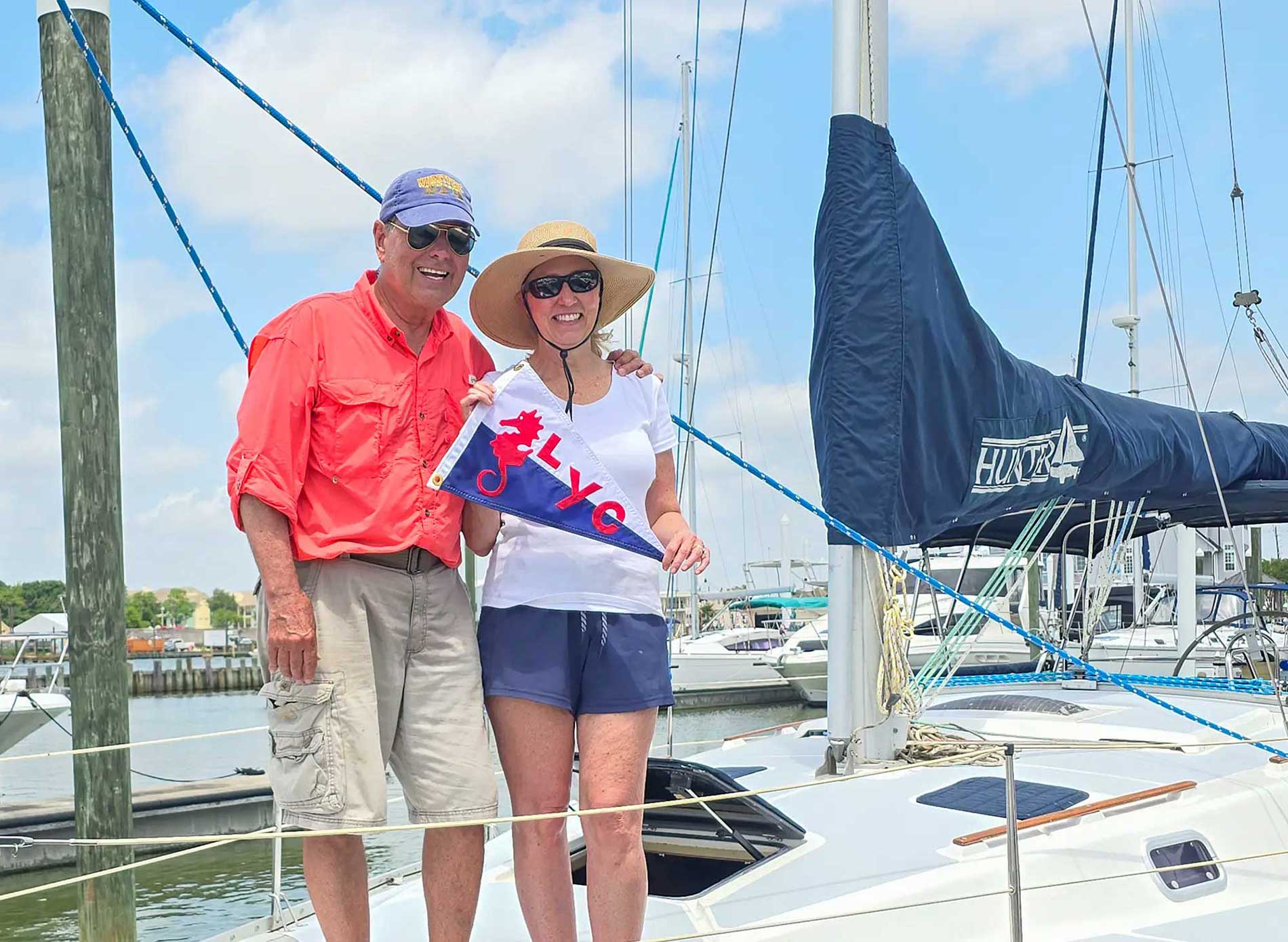 Smiling couple on a sailboat docked at a marina, holding a LYC burgee