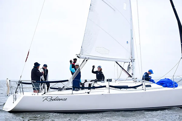 A group of five people on a white sailboat named Radiance, two waving, sailing on calm water under a cloudy sky.