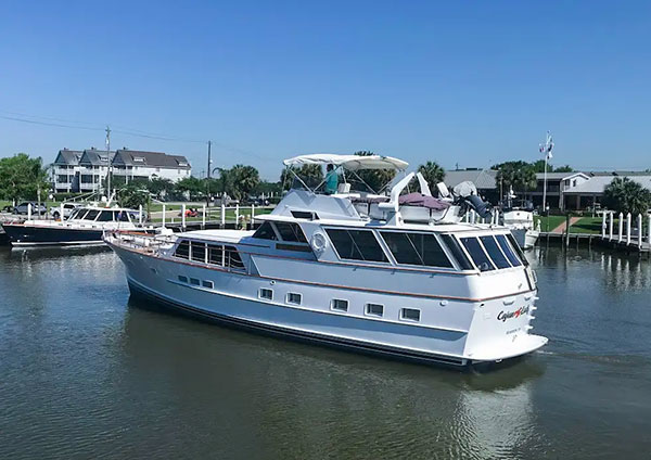 White luxury yacht docked in a calm harbor with houses and greenery in the background under a clear blue sky.