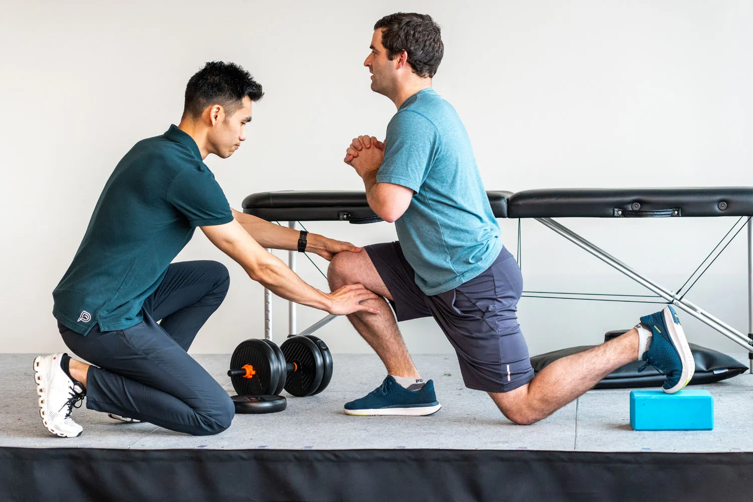 Physical therapist assisting a man doing a supported lunge stretch on a yoga block in a gym.