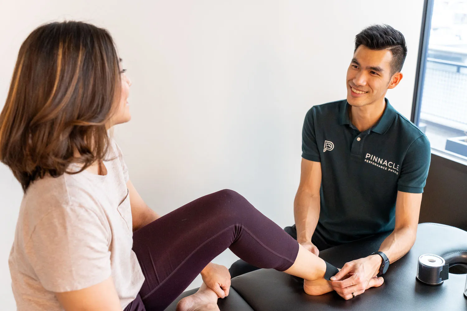 Physiotherapist in Pinnacle shirt examining a woman’s ankle on a treatment table, smiling.