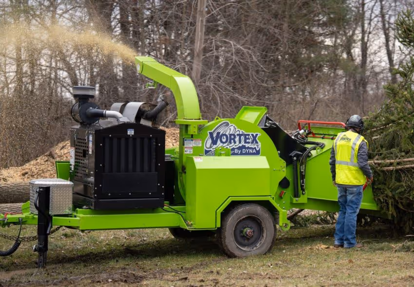 Worker operating a bright green Vortex wood chipper machine, shredding tree branches in an outdoor wooded area.
