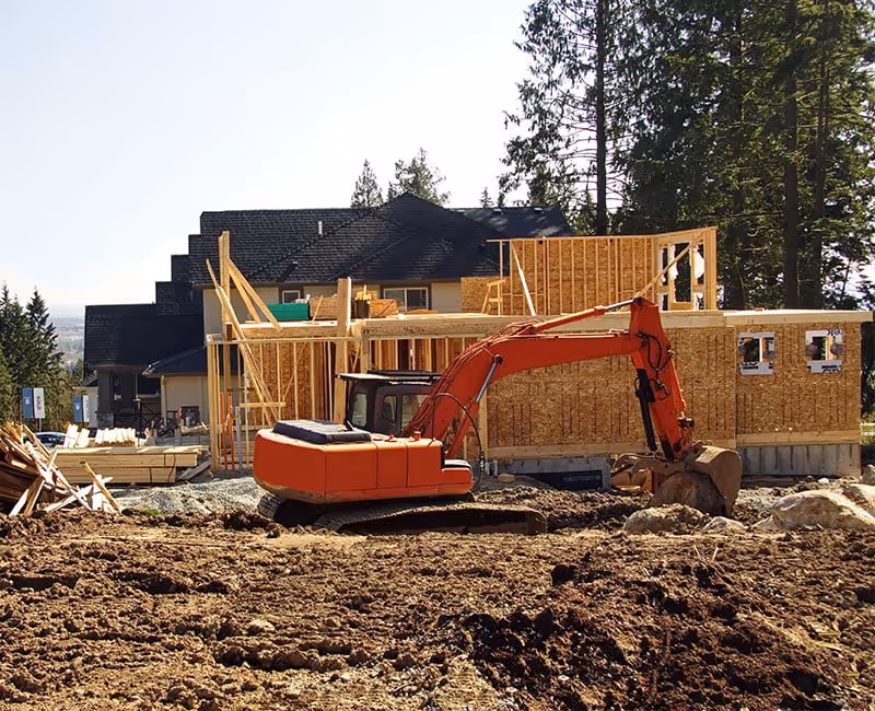 Orange excavator parked on dirt in front of a wooden frame house under construction surrounded by trees.