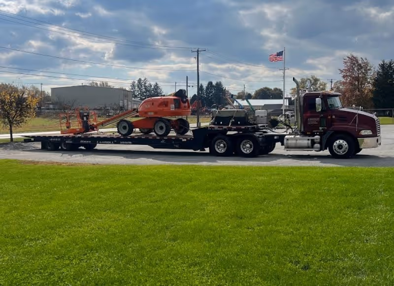 Maroon semi-truck with a flatbed trailer carrying a red aerial lift under a partly cloudy sky.