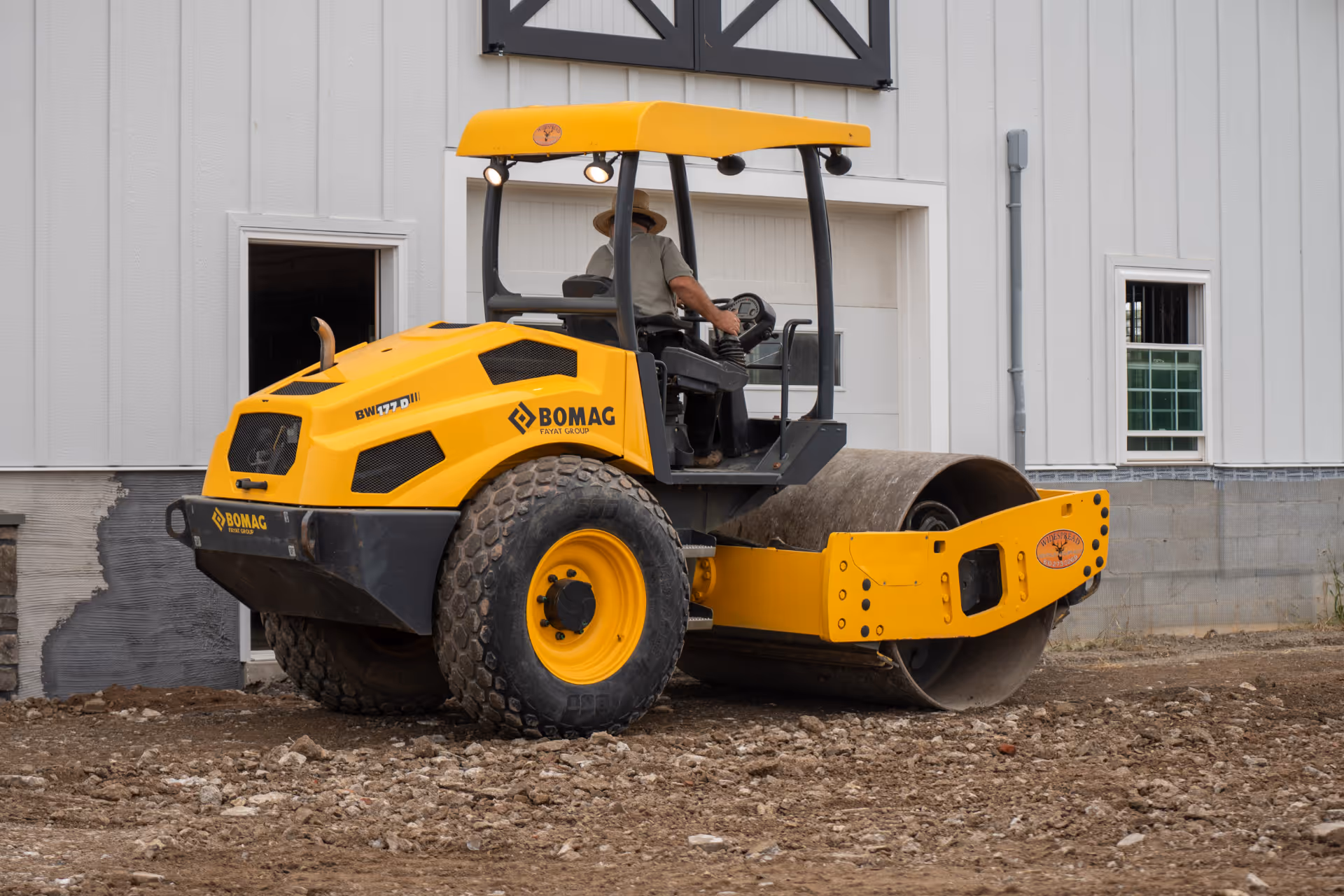Yellow BOMAG BW177D soil compactor operated by a man wearing a hat, parked on rough ground in front of a white building.