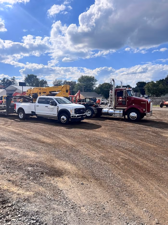White pickup truck and maroon semi-truck parked on a gravel lot under a partly cloudy blue sky.