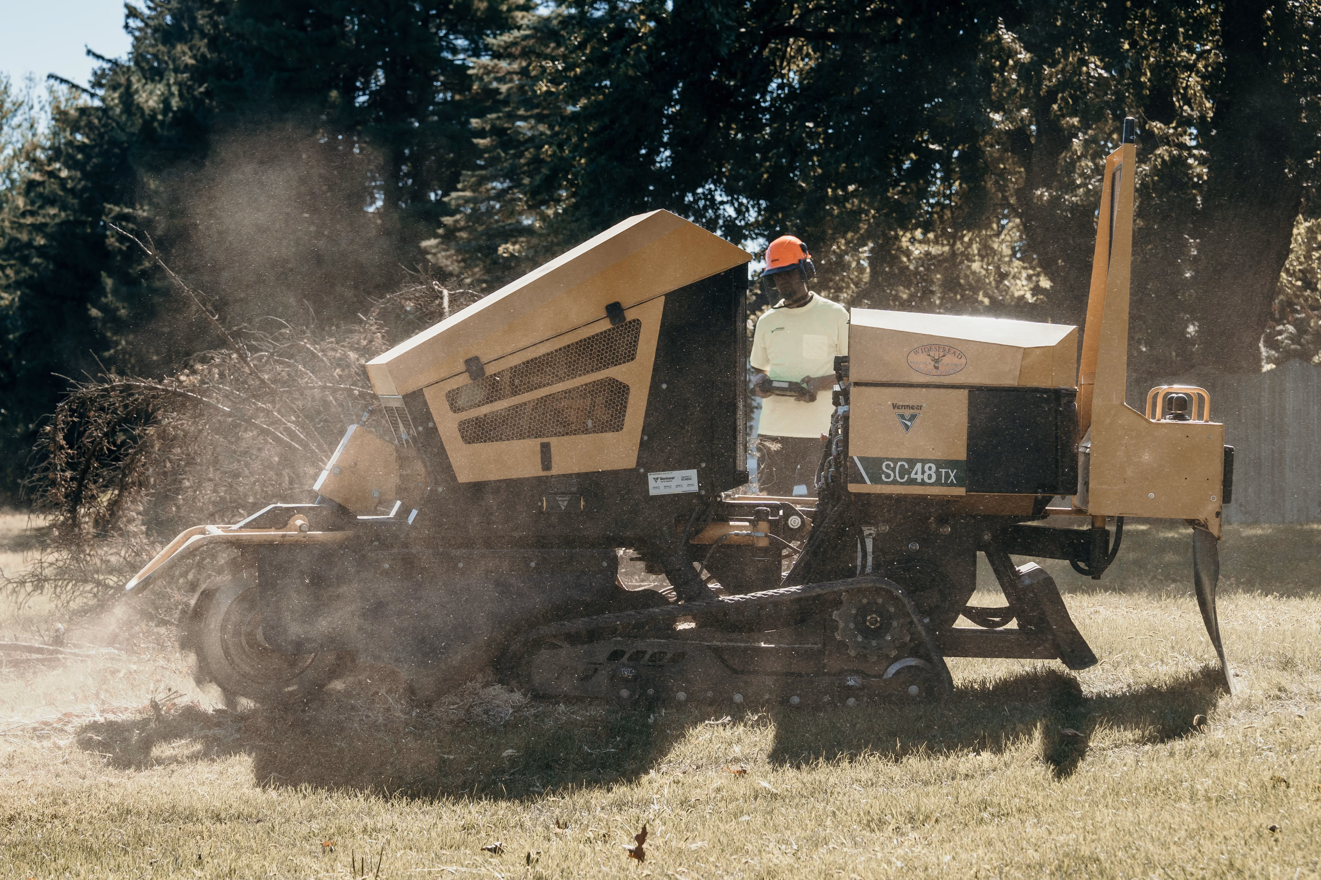 Worker in safety gear operating a Vermeer SC48TX stump grinder in a grassy area with wood chips and dust around.