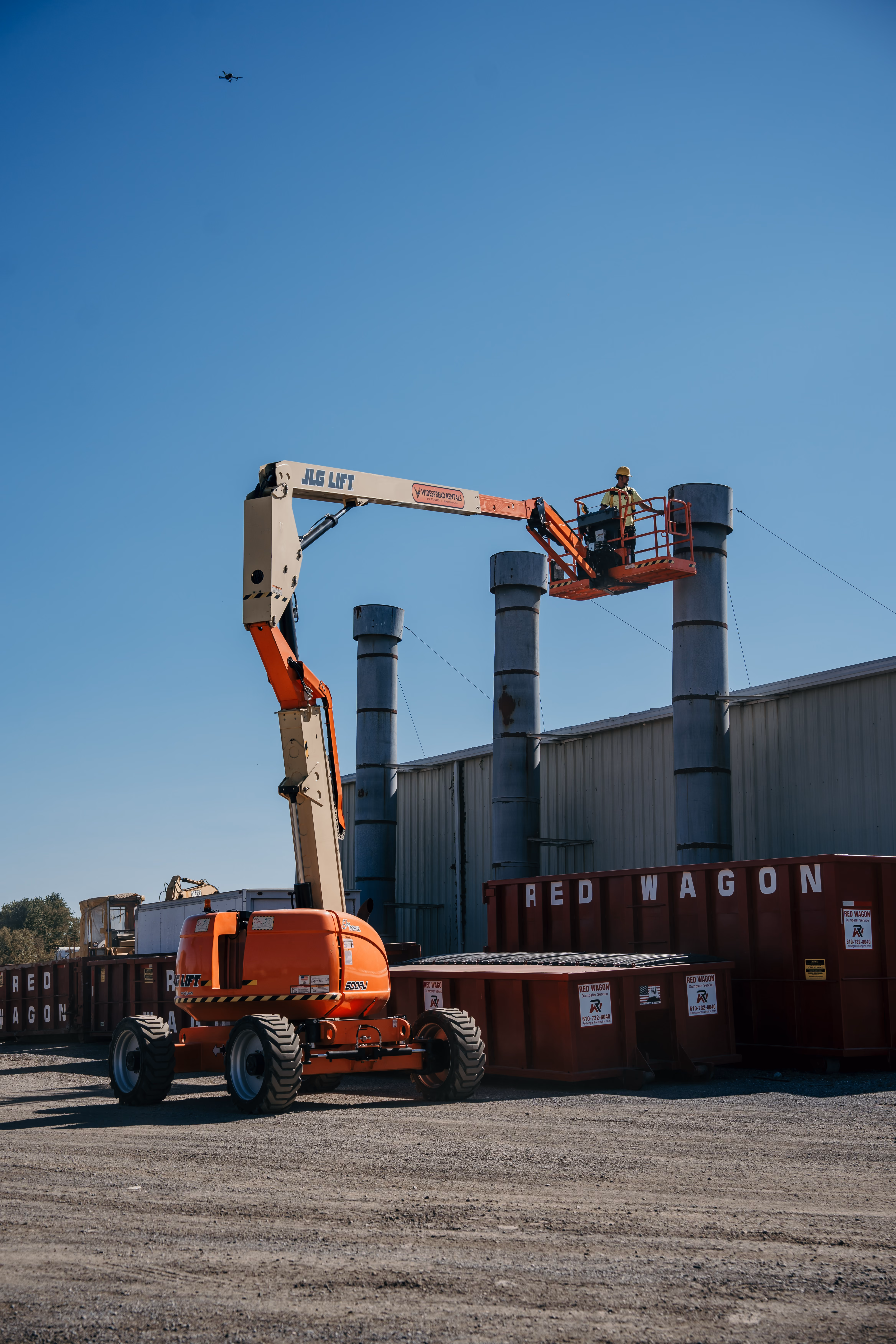 Worker in a yellow safety vest and helmet operating an orange JLG aerial lift near large metal chimneys and red dumpsters labeled RED WAGON.