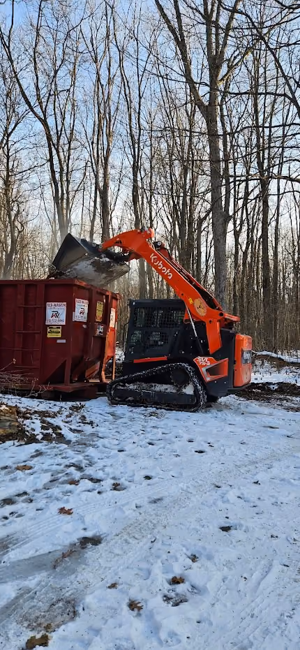 Kubota compact excavator loading debris into a red dumpster in a snowy wooded area.